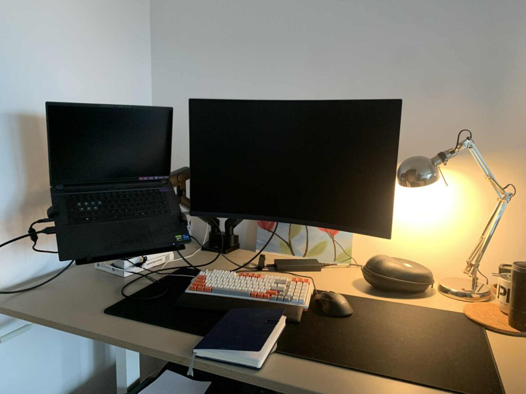 Minimalistyczne stanowisko pracy z jasnym biurkiem: po lewej laptop na podstawce, pośrodku duży zakrzywiony monitor, przed nimi klawiatura mechaniczna i mysz. Po prawej zapalona metalowa lampka biurkowa, etui na okulary i kubek. Na blacie leży także notes, widoczne są kable oraz mata na biurko, całość ustawiona przy białej ścianie.